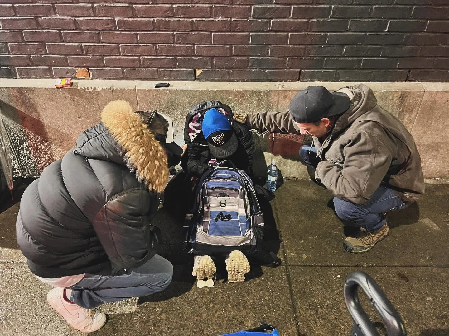 Two outreach volunteers kneeling to pray with a man on a downtown sidewalk at night